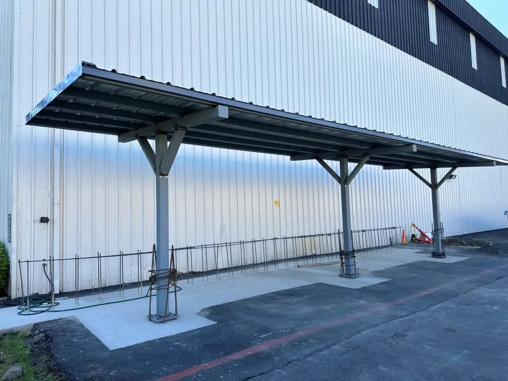 A gray metal carport structure with three support posts installed along the side of a white corrugated industrial building.