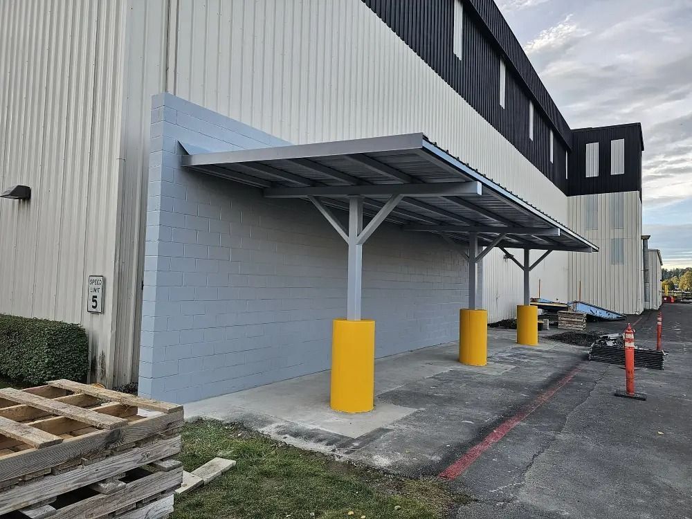 A grey metal awning attached to a light-colored building wall, supported by three columns with yellow protective guards.