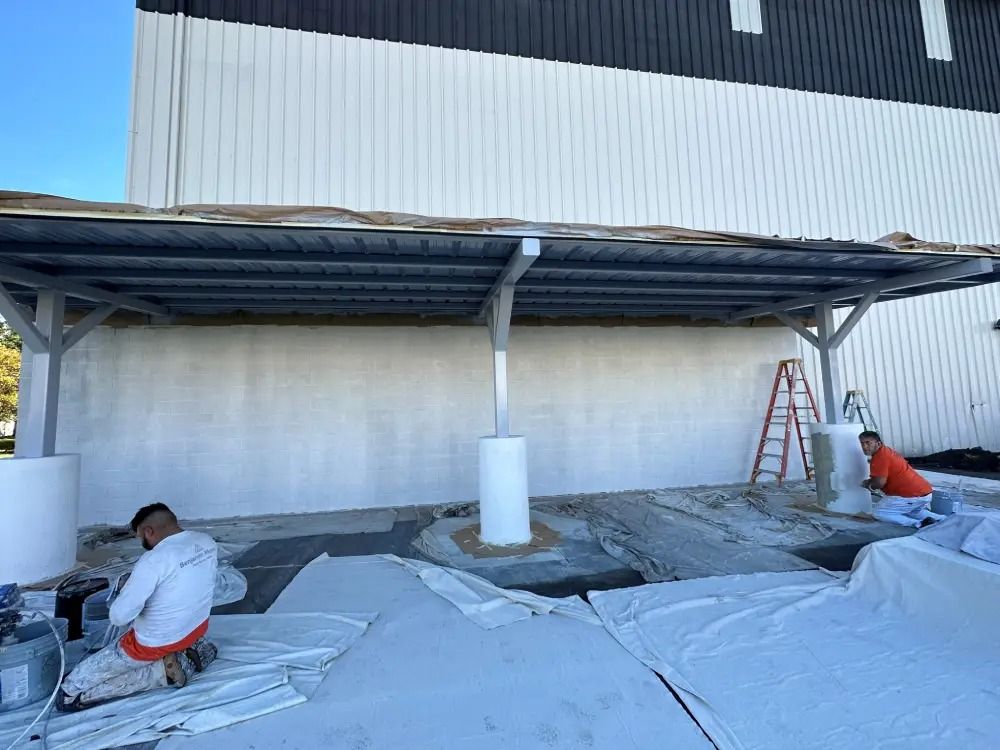 Two workers paint the exterior wall of a building white, beneath a metal canopy structure.