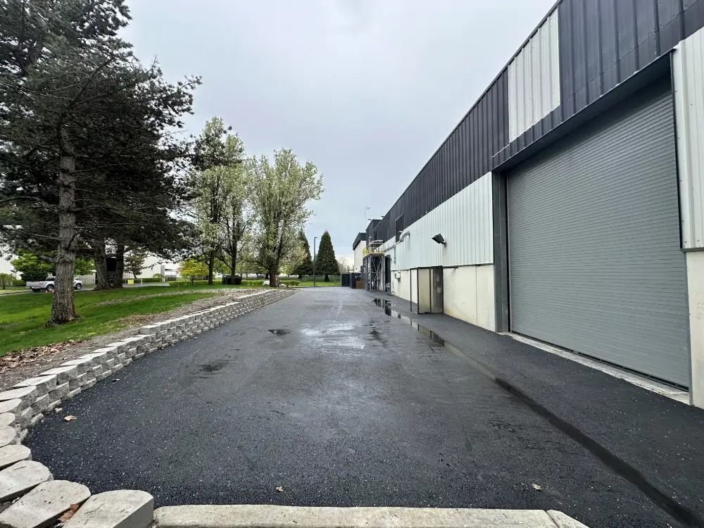 A paved driveway leads toward a commercial building with a large gray roll-up door alongside a grassy area with trees.