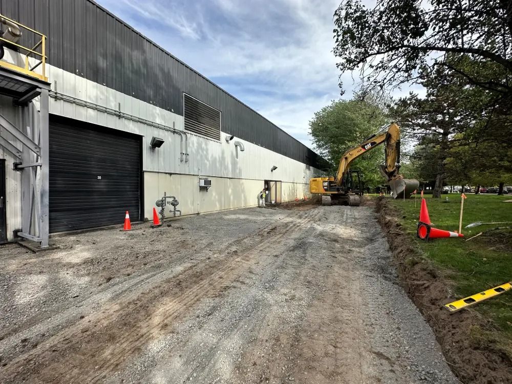 An excavator parked on a gravel construction site beside a large, industrial warehouse building with orange safety cones.