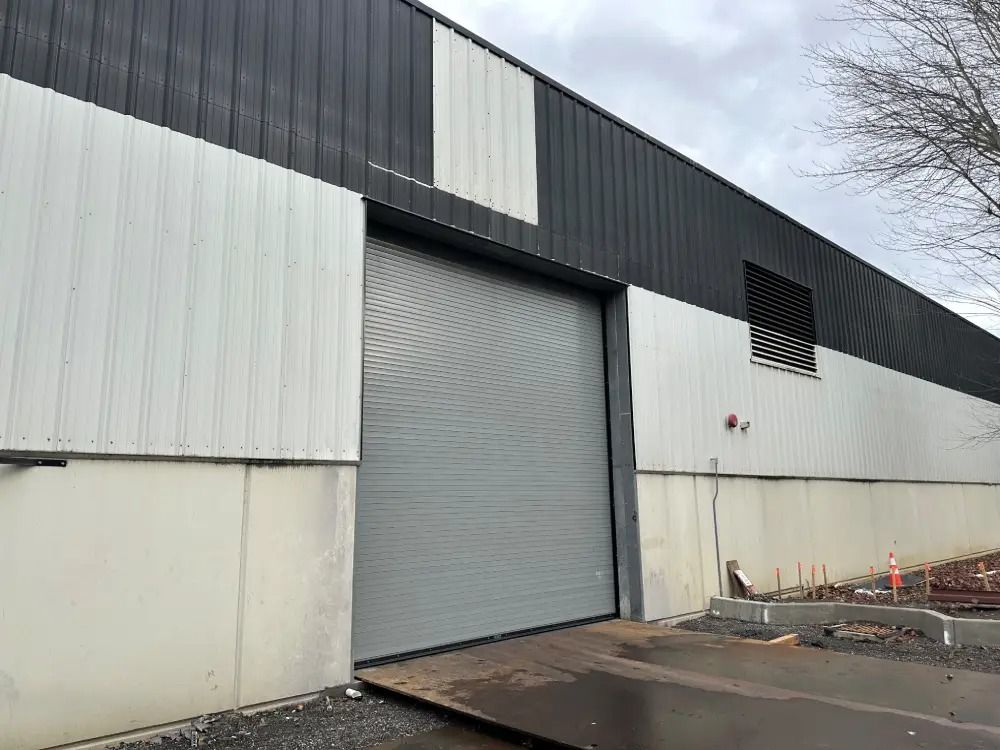 An industrial warehouse exterior with a large, gray roll-up metal door, white lower walls, and dark corrugated siding.