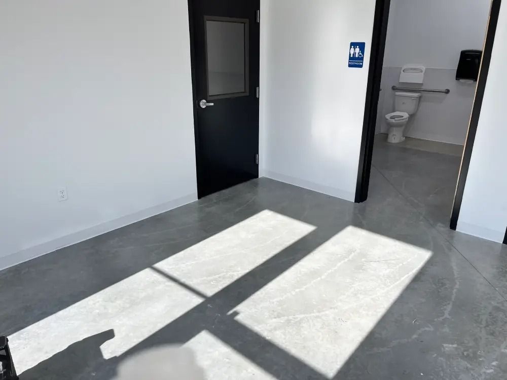 A black door stands next to a restroom entrance featuring a wheelchair-accessible toilet, with sunlight on a gray floor.