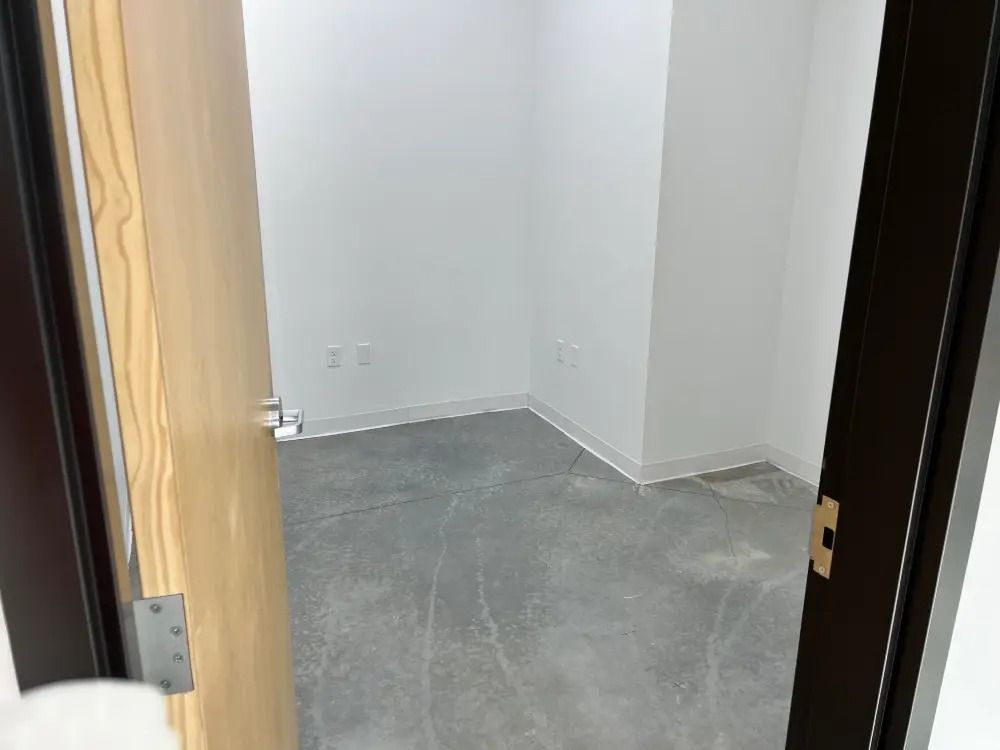 An open wooden door reveals a small, empty room with plain white walls and a grey concrete floor.
