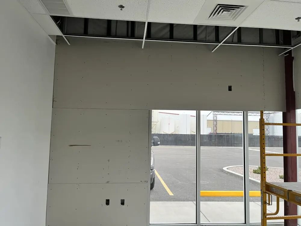 Interior view of a construction site showing unfinished drywall panels on a wall next to large windows and a scaffold.