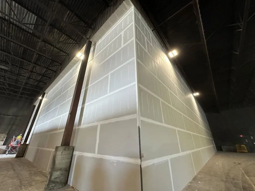 A corner of an unfinished drywall structure in a large, dark warehouse space with exposed ceiling beams and metal pillars.