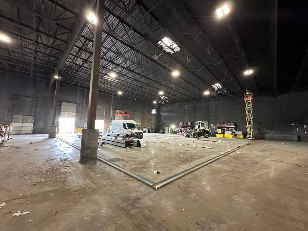 An empty, dimly lit industrial warehouse with high ceilings, concrete floors, and scattered construction equipment.