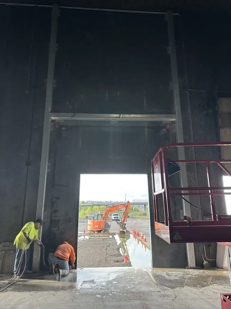 Two workers in high-visibility gear work near a large, open doorway in a dimly lit, unfinished industrial space.