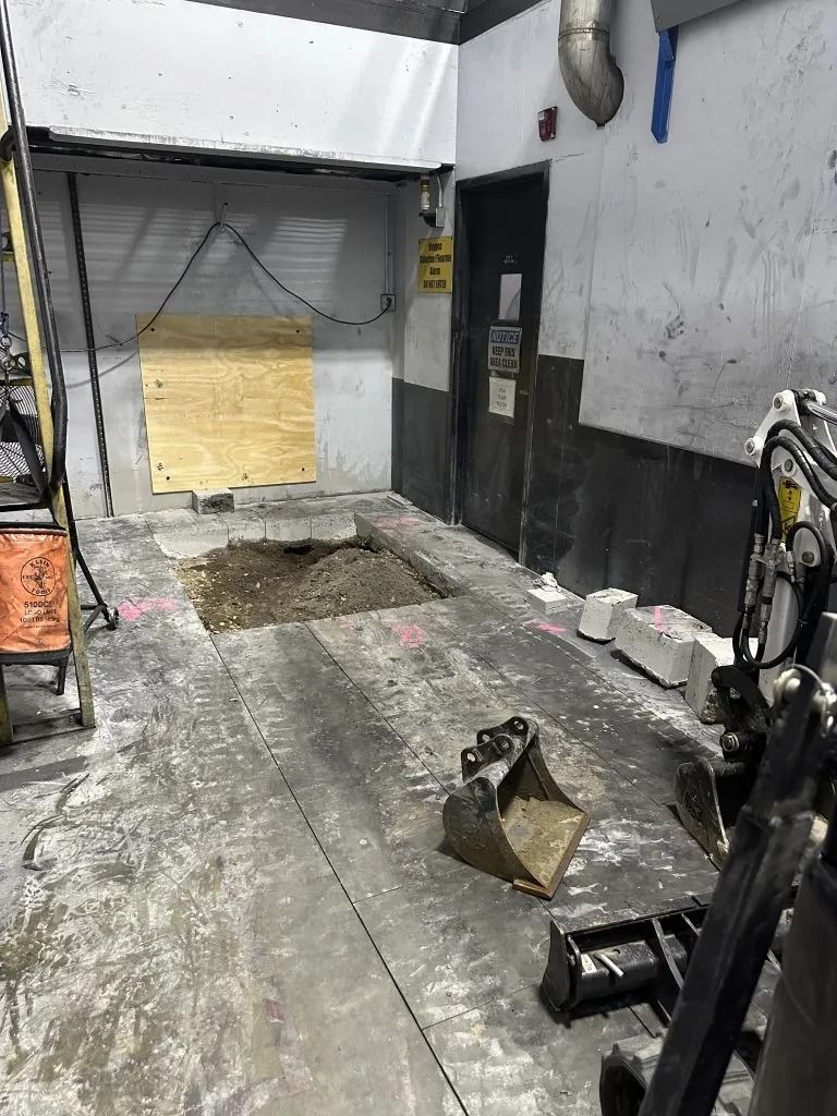 An indoor construction site shows a rectangular hole in a concrete floor, a plywood-covered opening, and excavator parts.