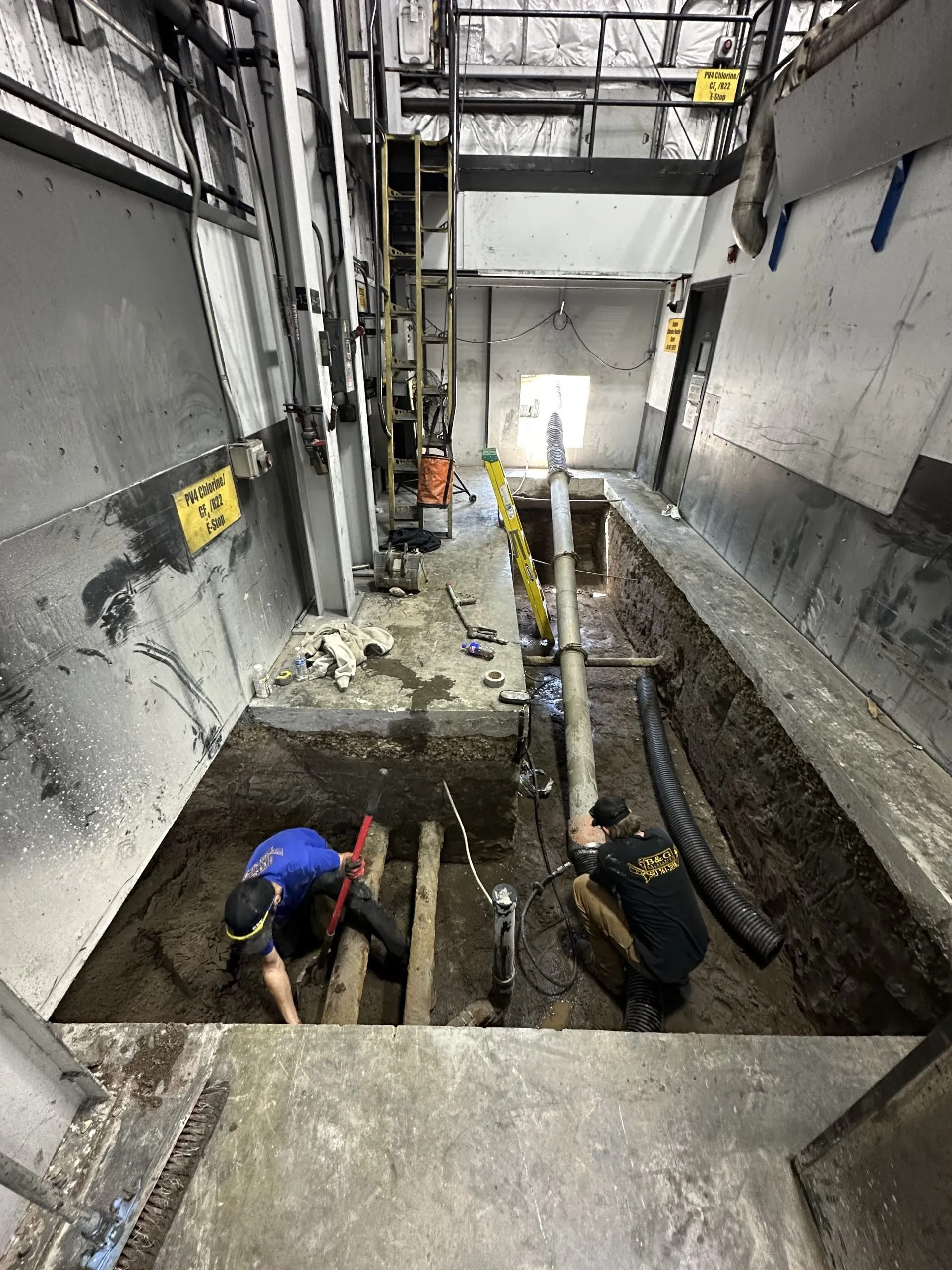 Two workers in an underground trench repair pipes within a concrete construction site.