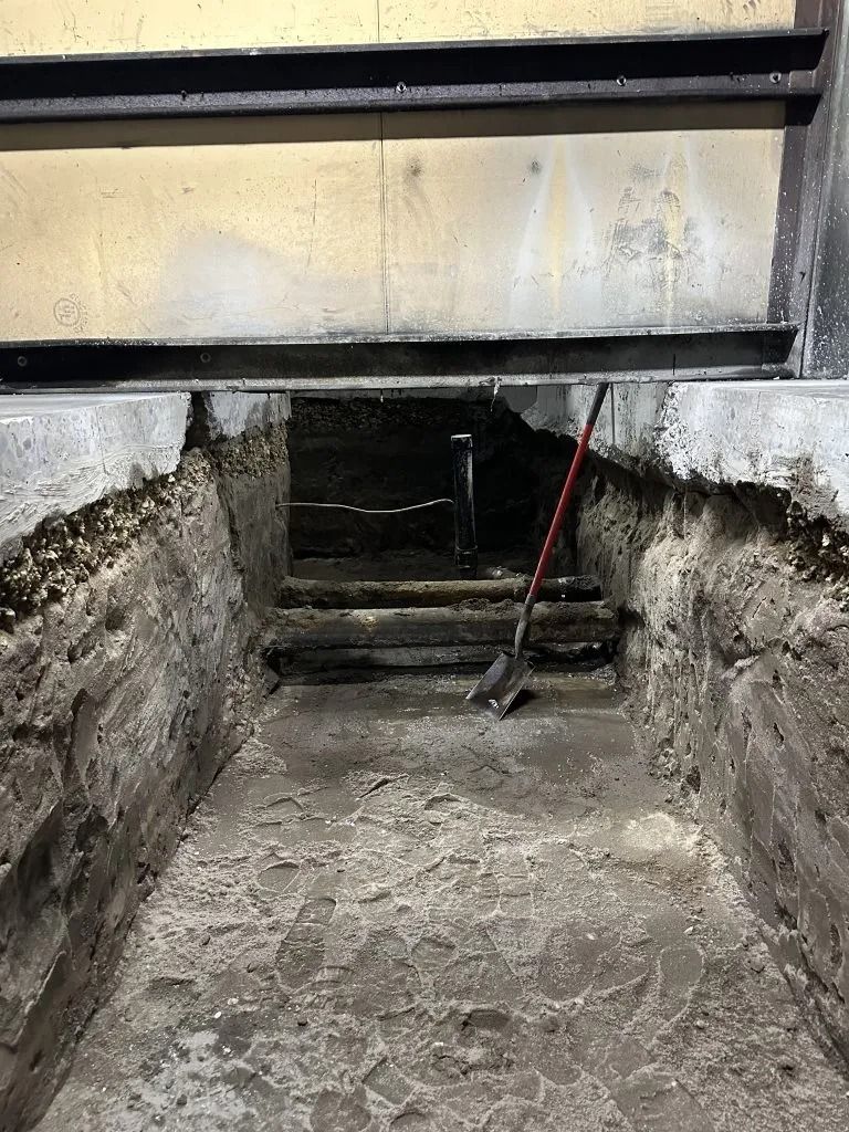 A trench dug into a concrete floor leads to a dark, recessed doorway, with a shovel resting against the back wall.