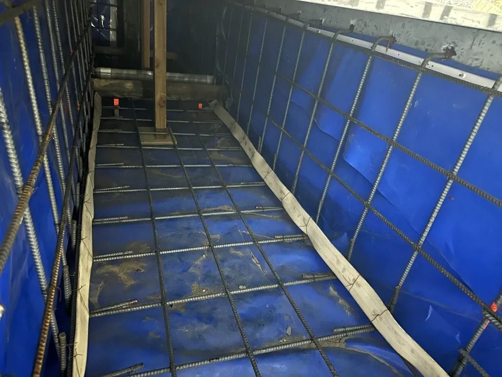 Construction site showing a trench or foundation formwork lined with blue plastic, reinforced with a steel rebar grid.