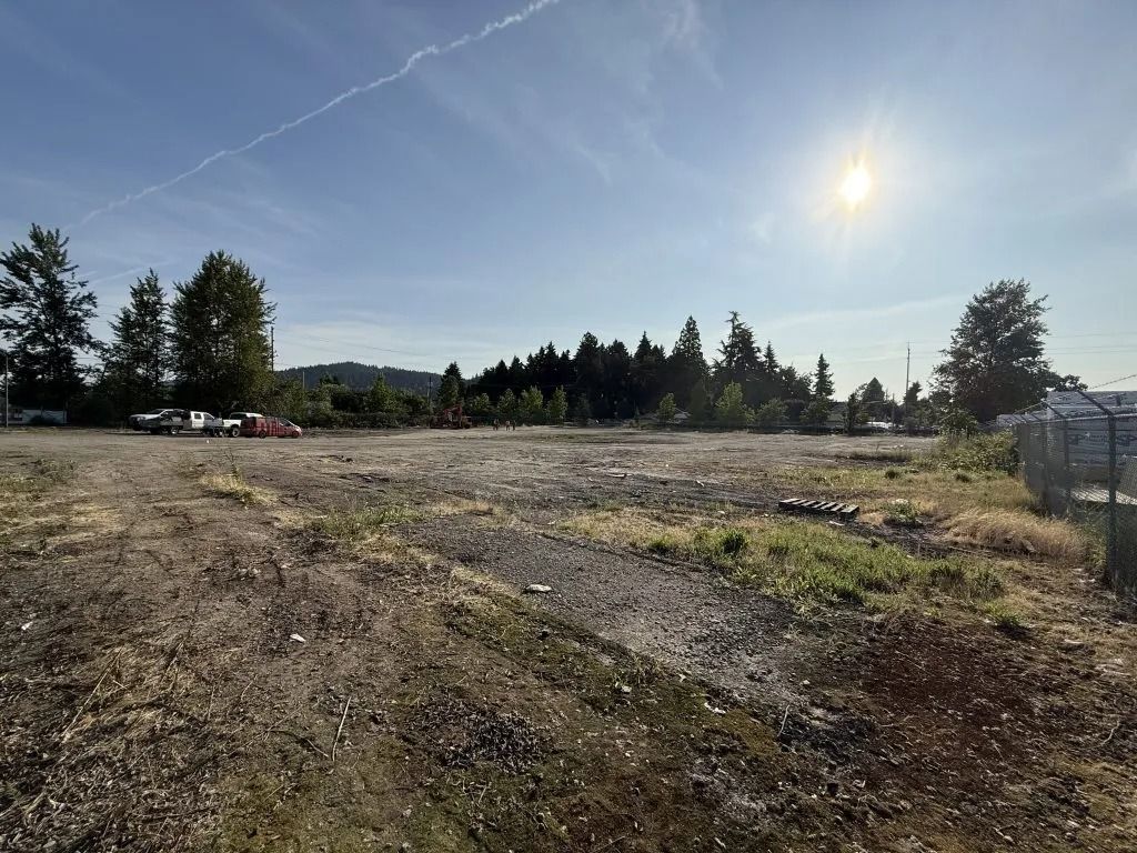 A flat, open, dirt-covered lot with scattered vegetation under a sunny blue sky, framed by trees in the background.