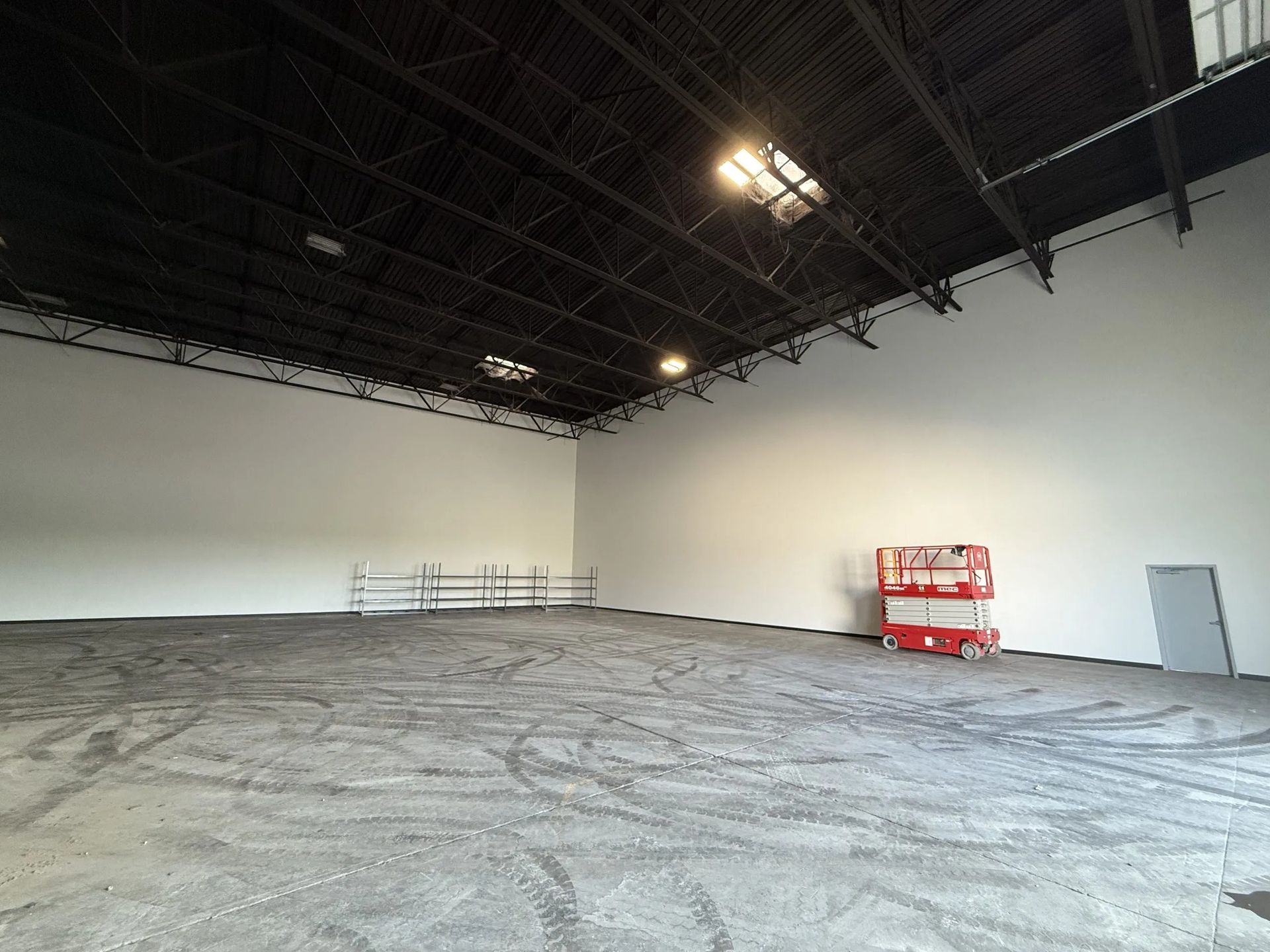 Empty industrial warehouse with high black ceilings, white walls, concrete floors with tire marks, and a red scissor lift.