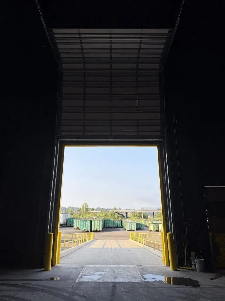 An open warehouse bay door looking out to an exterior yard with rows of green shipping containers under a clear sky.