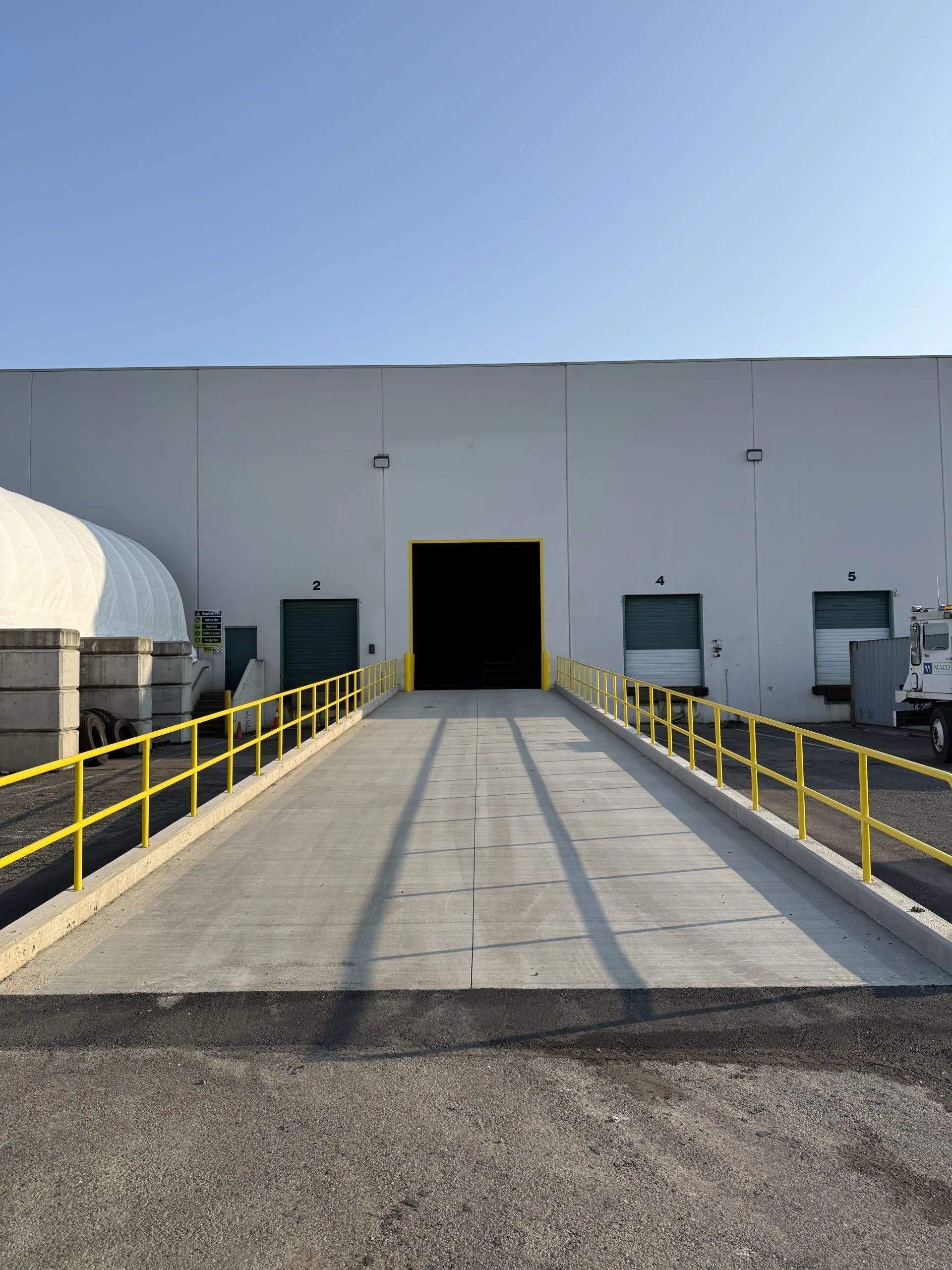 A concrete loading dock ramp with yellow railings leading to an open warehouse doorway on a white building.