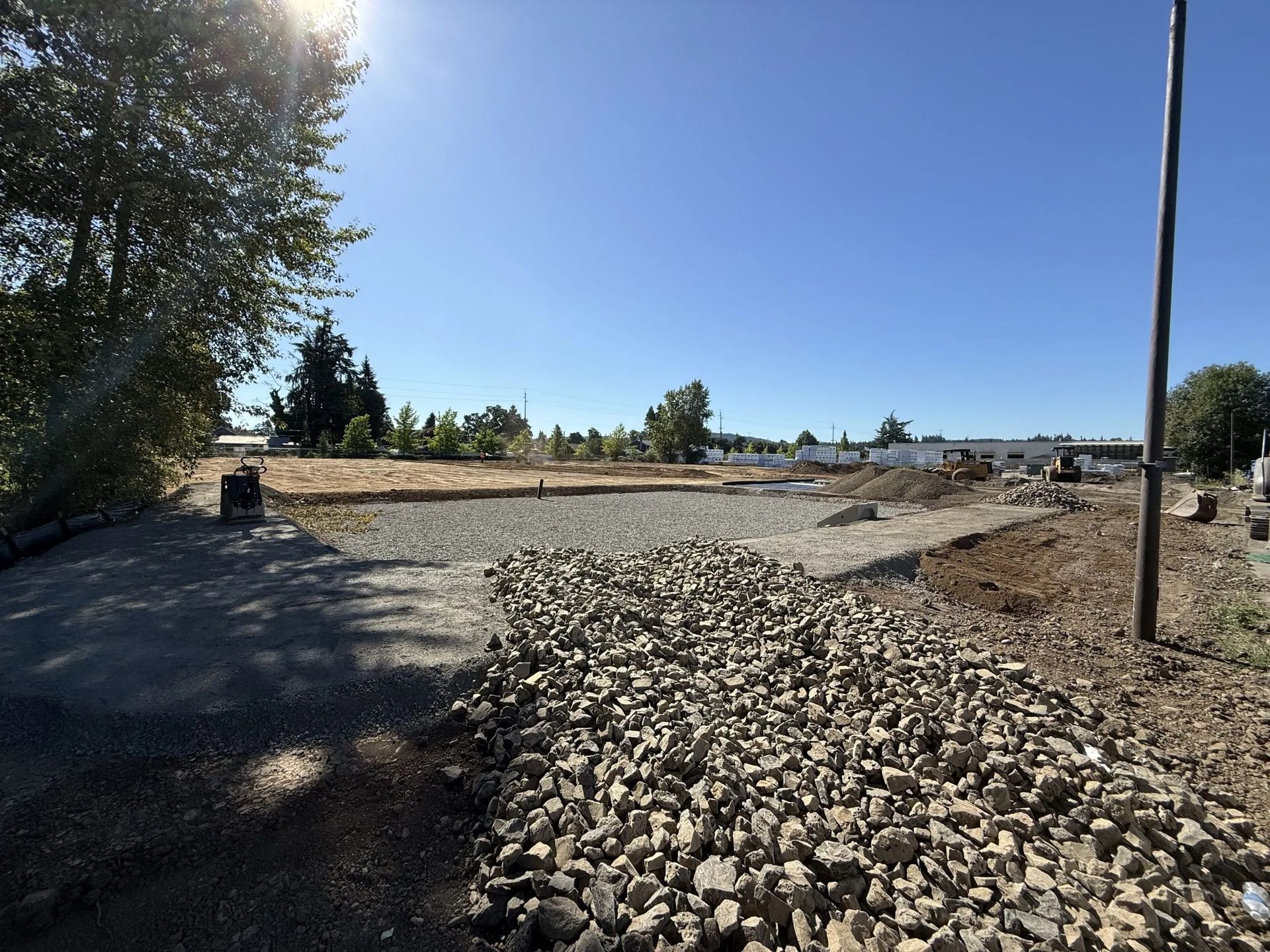A gravel-covered construction site under a bright blue sky, with trees on the left and a utility pole on the right.