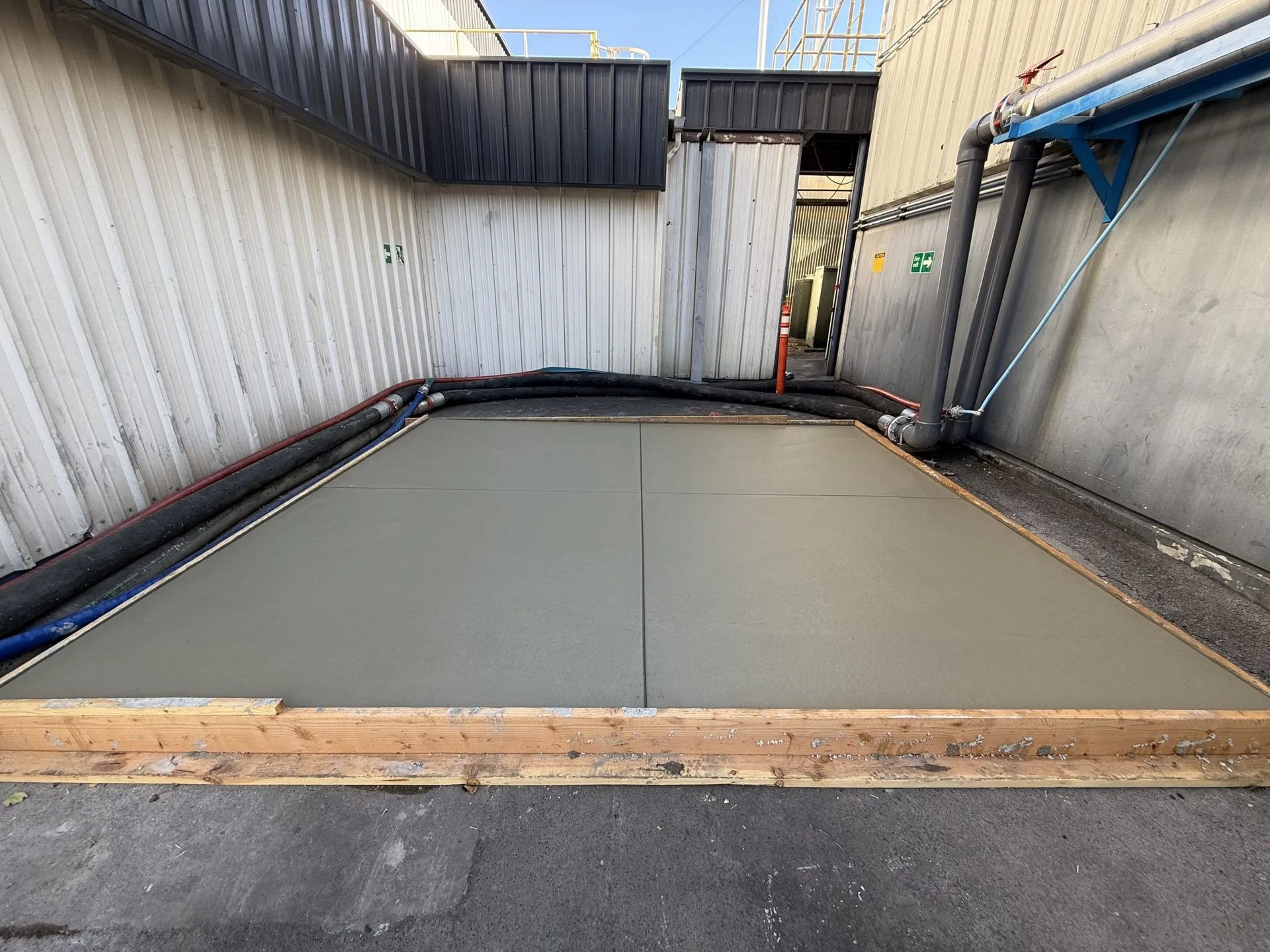 A freshly poured concrete pad framed with wooden boards between two industrial building walls.