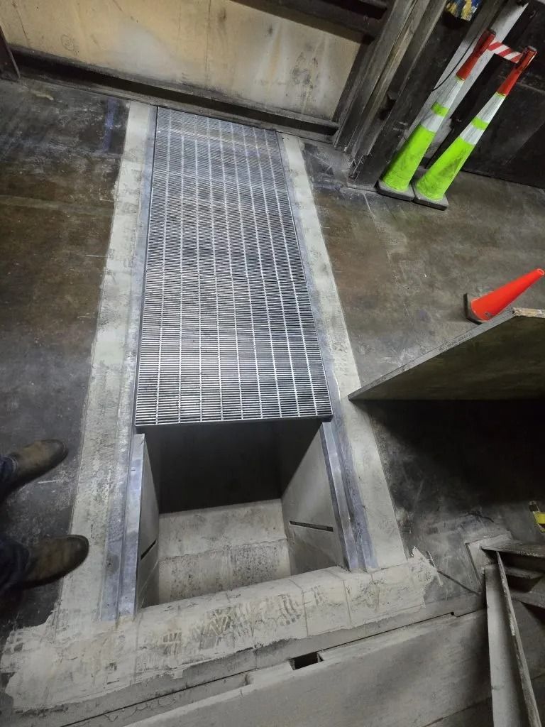 A metal grate partially covers an open rectangular trench in a concrete floor, with safety cones nearby.