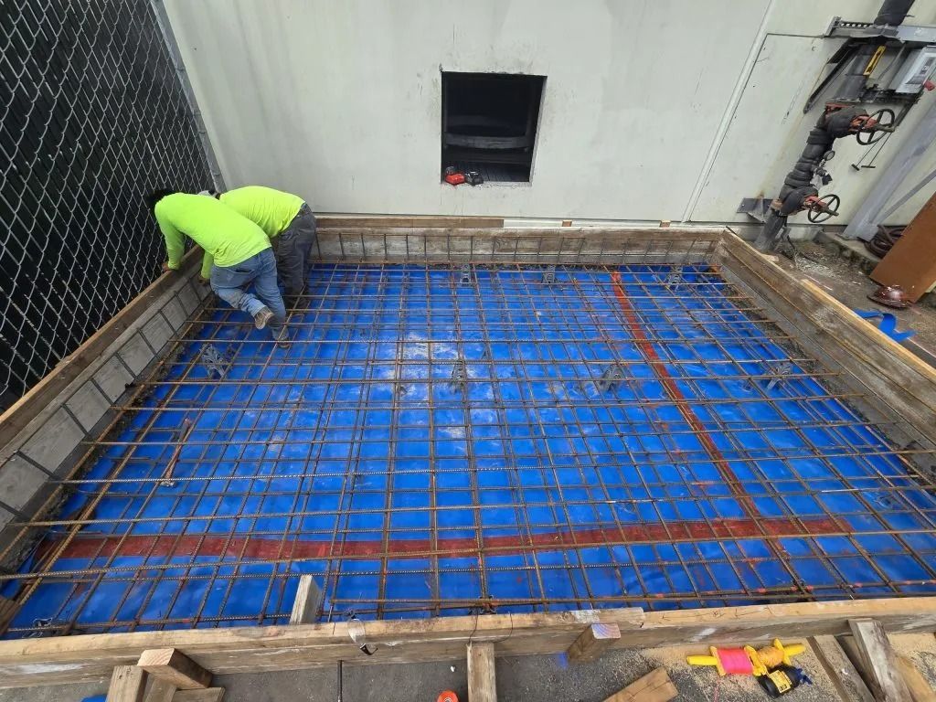 Two construction workers in neon shirts build a rebar grid over a blue vapor barrier inside a wooden concrete form.
