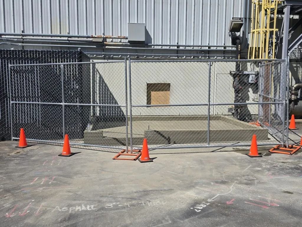 A fenced-off construction area with a concrete slab in front of a building wall, surrounded by several orange traffic cones.