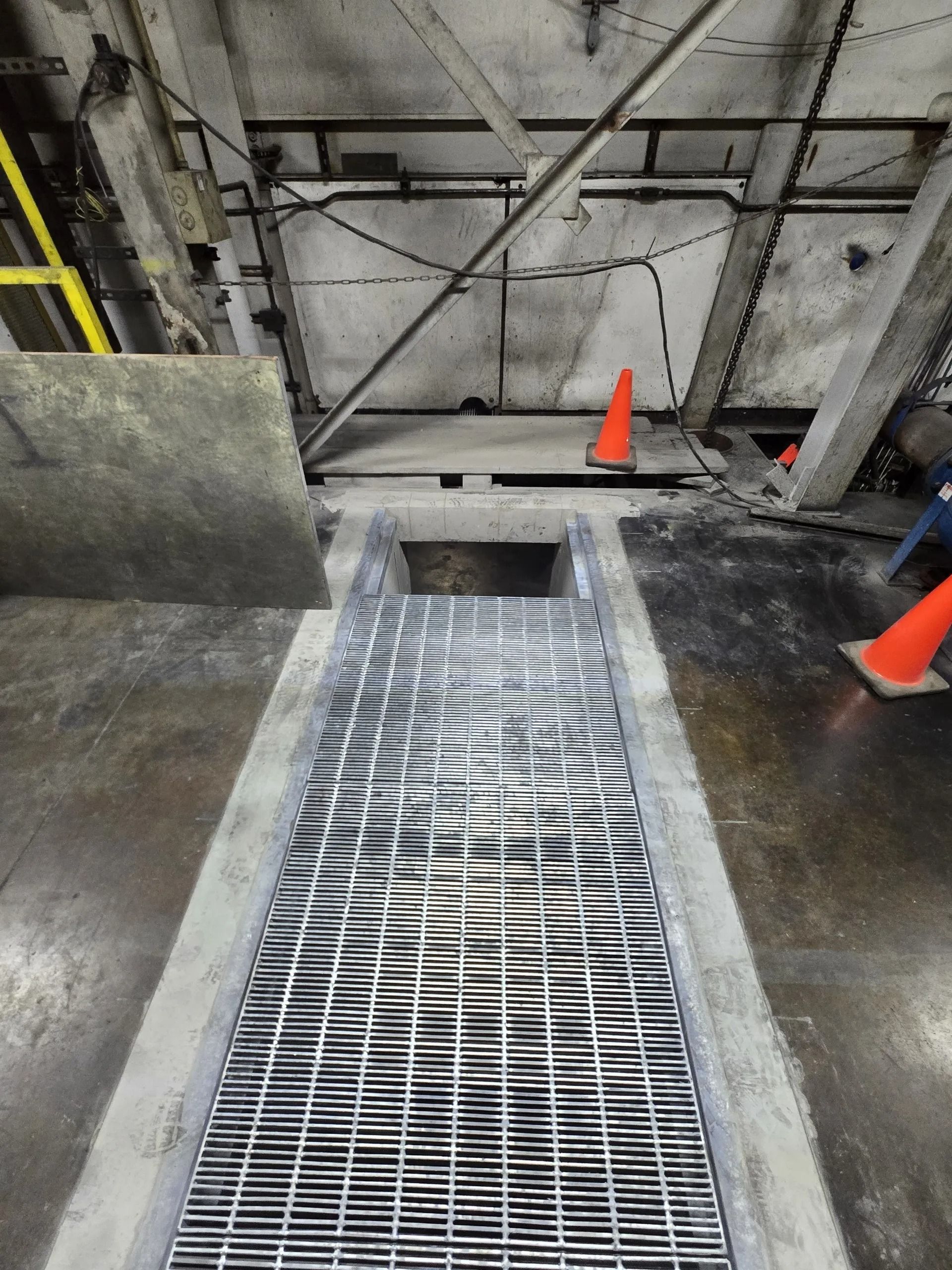 A metal grate walkway leading to a dark opening in a gray, industrial concrete space with orange safety cones.