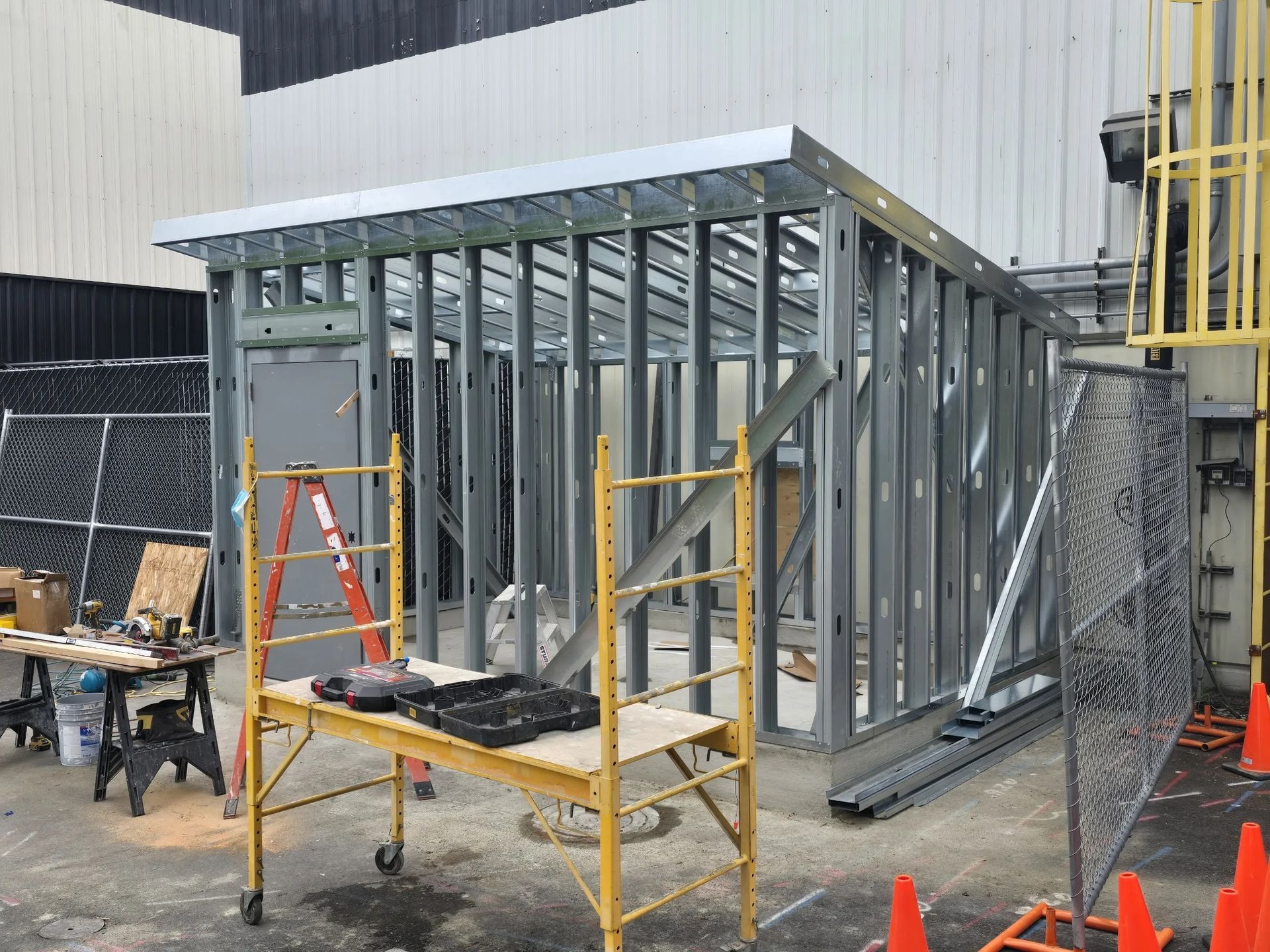 Construction of a metal-framed structure with a sloped roof at a building site, featuring yellow scaffolding in the front.