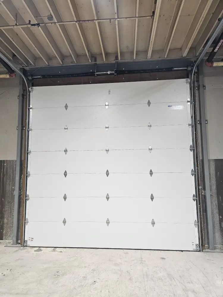 A closed, white industrial roll-up door installed in a concrete warehouse space with an exposed ceiling.