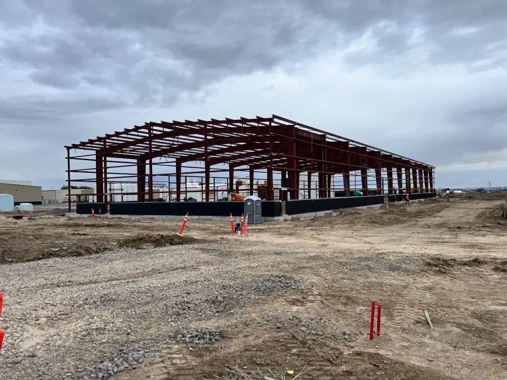 The skeletal red-steel frame of a large industrial building under construction on a dirt lot under a cloudy sky.
