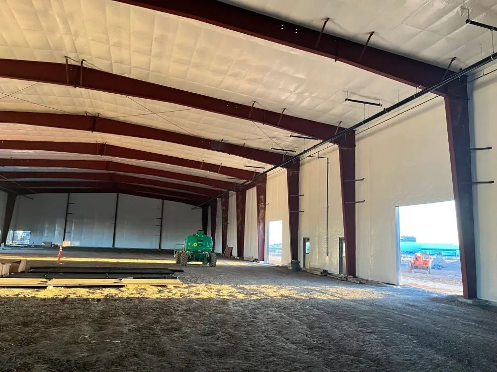 Interior view of a large, unfinished industrial warehouse with steel framing, white walls, and a dirt floor.