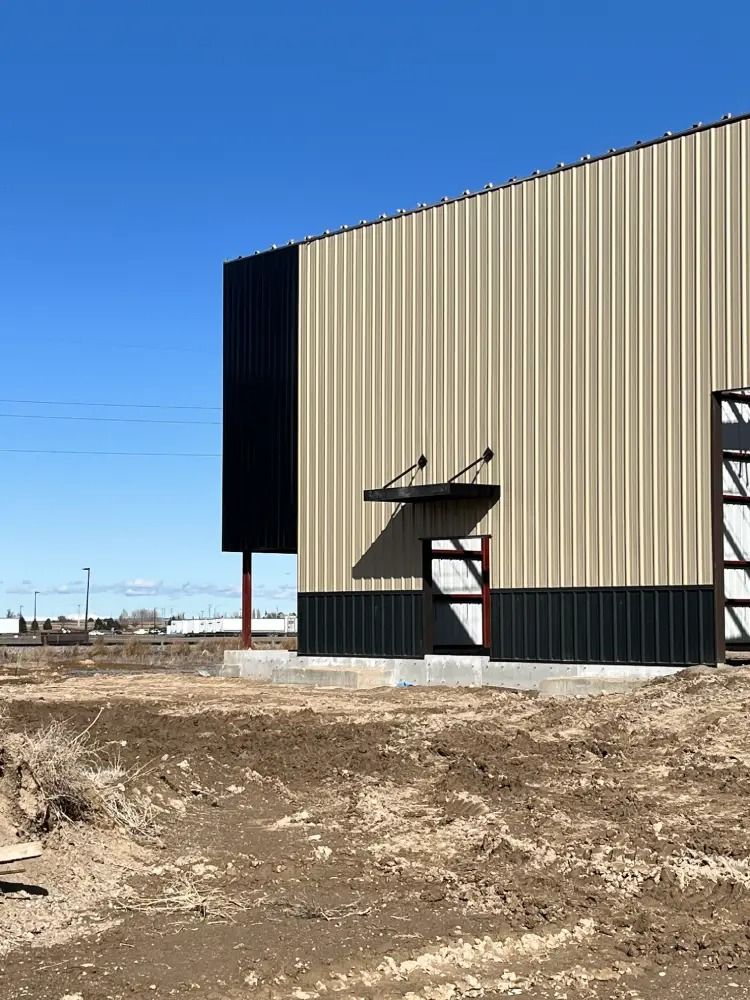 A metal building exterior with light tan and black vertical panels, a small doorway, and a metal awning under a blue sky.
