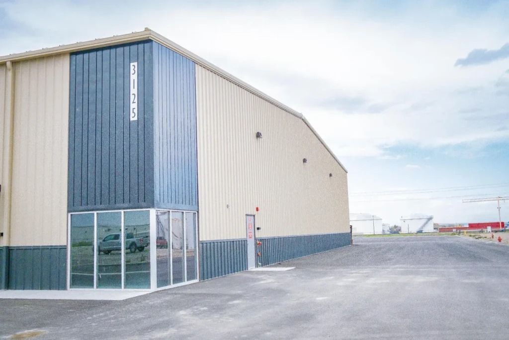 A tan and dark-gray industrial warehouse with glass windows on a gravel lot under a cloudy sky.