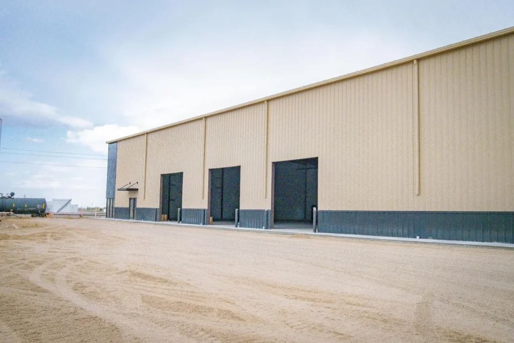 A large, tan, corrugated metal warehouse building with three dark roll-up doors and gray trim, set in a dry, open lot.