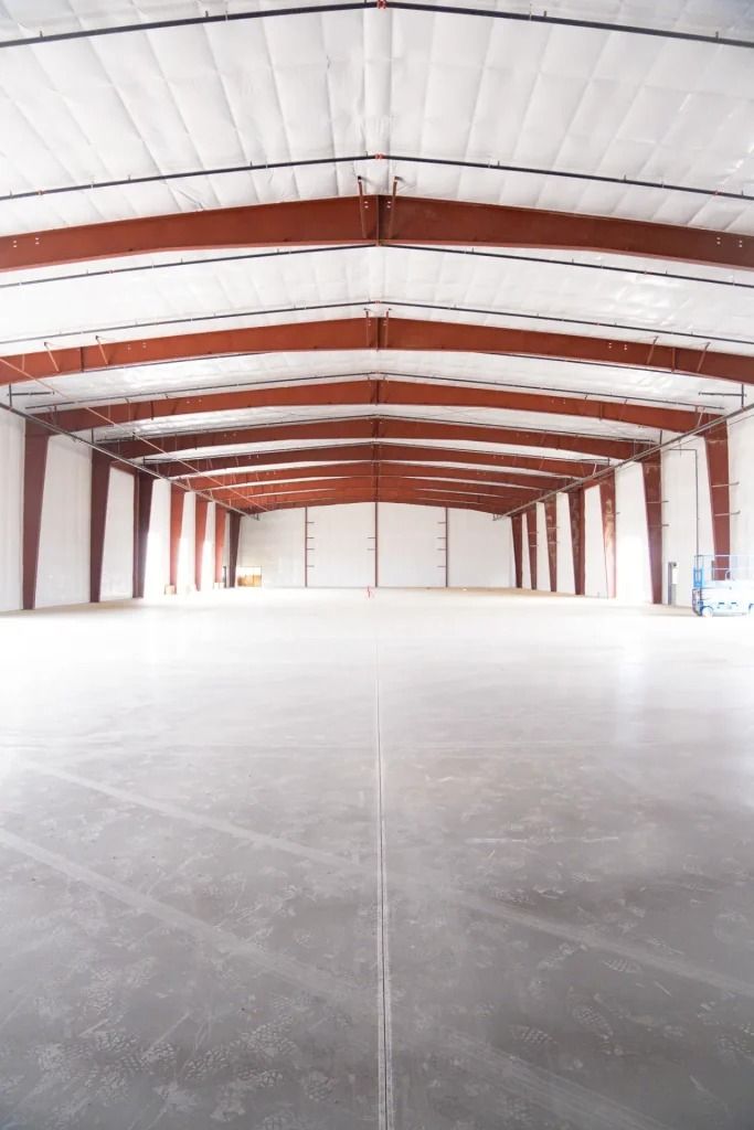 A large, empty industrial warehouse with white walls, a smooth concrete floor, and red steel support beams.