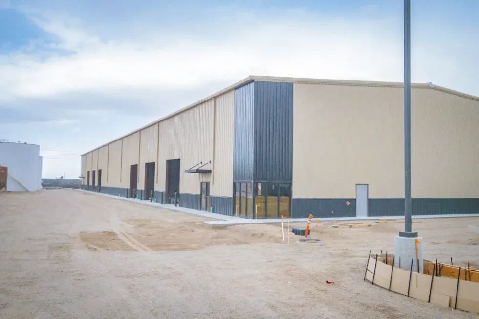 A large, beige warehouse building with dark gray paneling, under-construction gravel grounds, and a tall light pole.