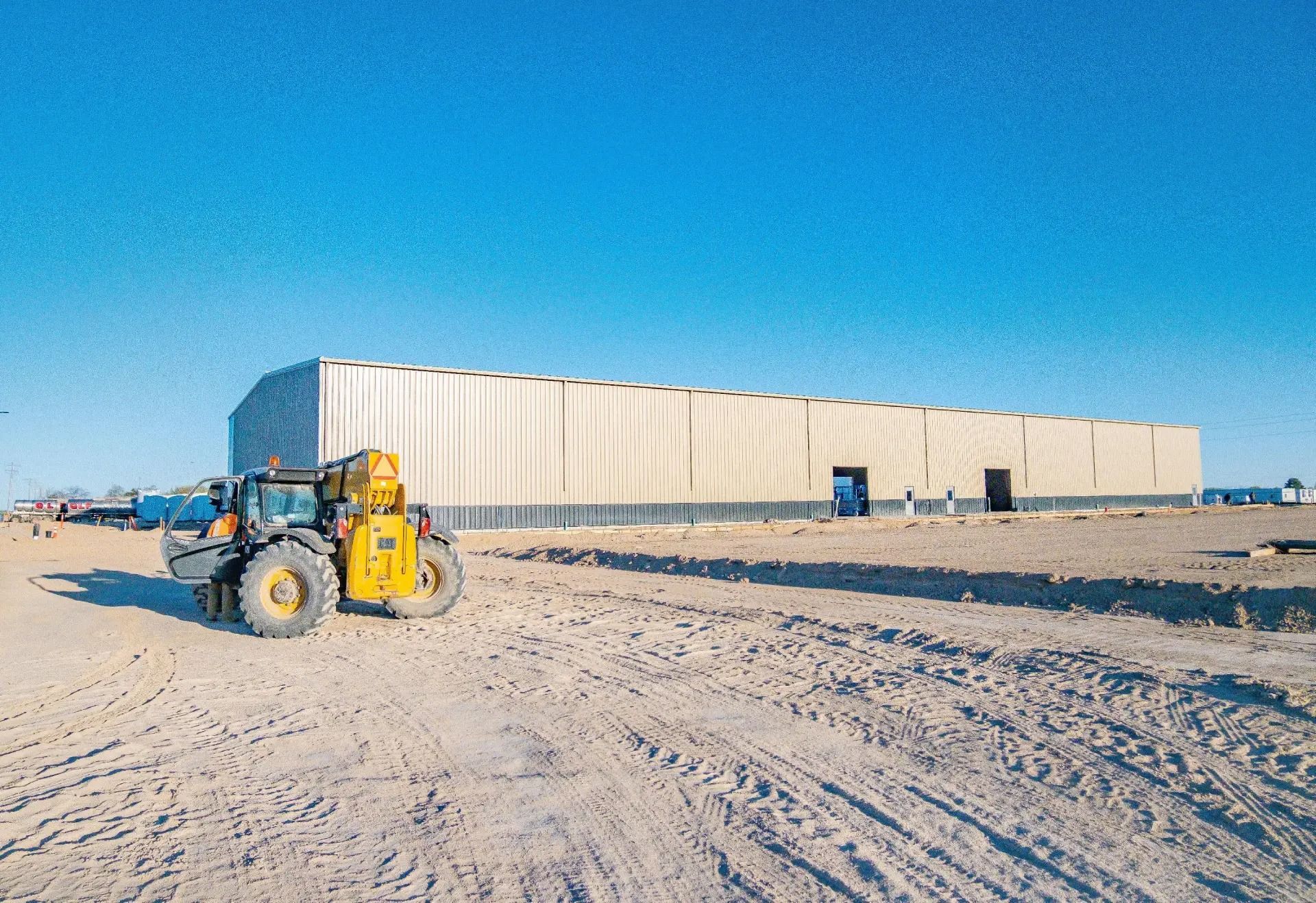 A yellow telescopic forklift parked on a dirt construction site in front of a large, long metal warehouse building.