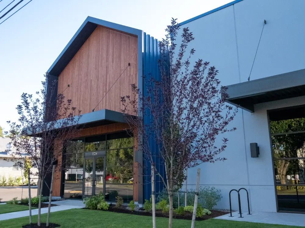 Modern building entrance with a wooden gabled facade, blue metal accents, and thin trees planted on a grassy lawn.