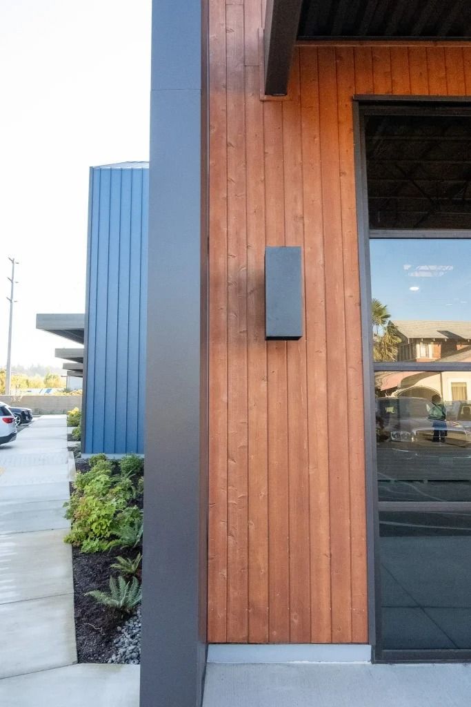 Modern building exterior featuring vertical wood paneling, a dark gray corner frame, and a minimalist wall sconce.