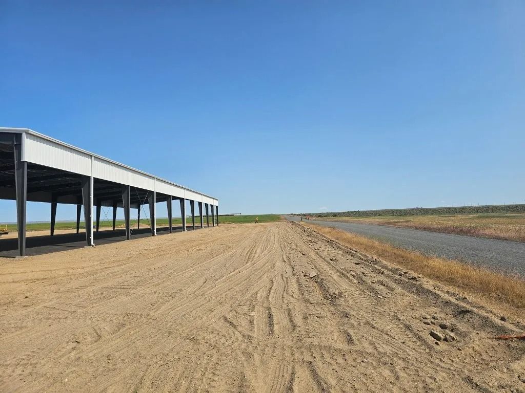 A large, open-sided metal warehouse structure stands beside a gravel road under a clear blue sky.