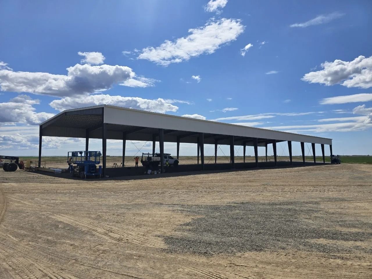 A long, open-sided steel structure under construction on a dirt lot under a bright blue sky with scattered clouds.