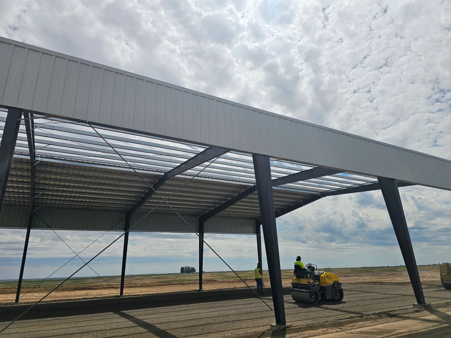 A large, open-air metal building structure with a worker operating a yellow roller on the dirt floor underneath.