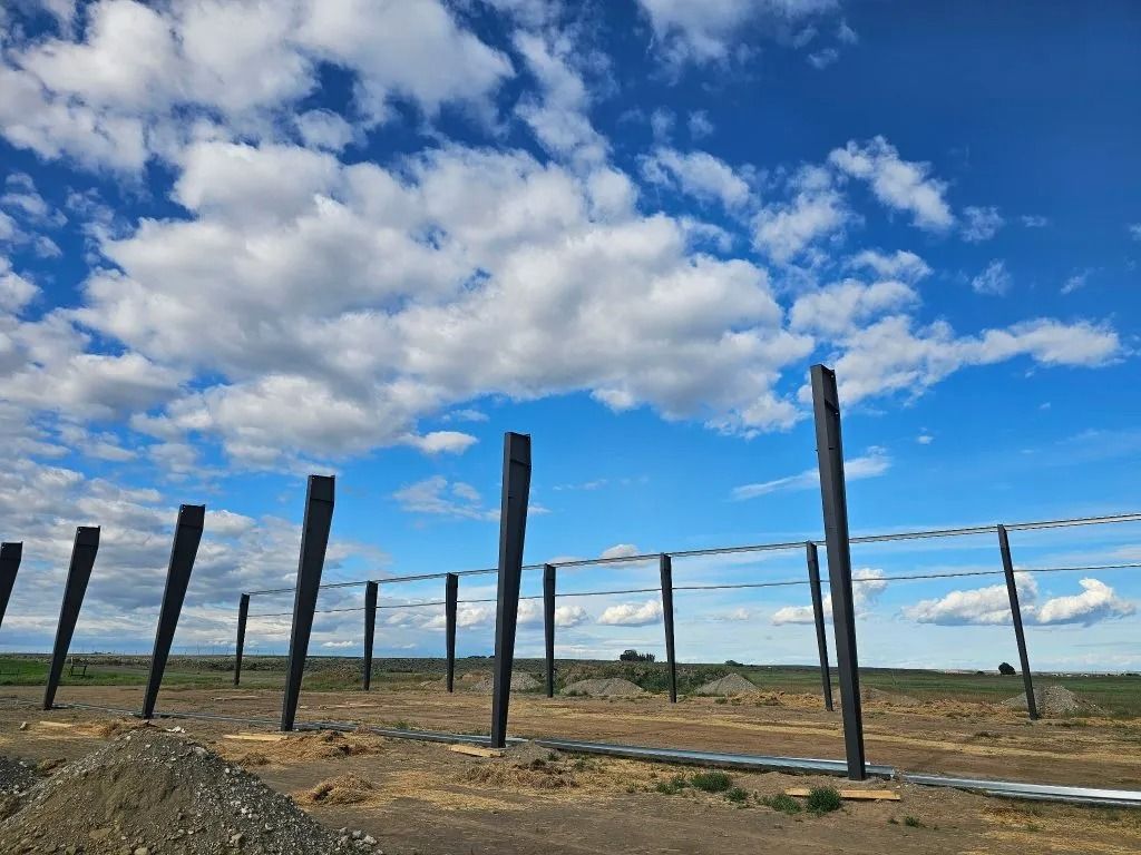 A row of dark metal support beams stands on a dirt construction site under a bright blue sky with scattered clouds.