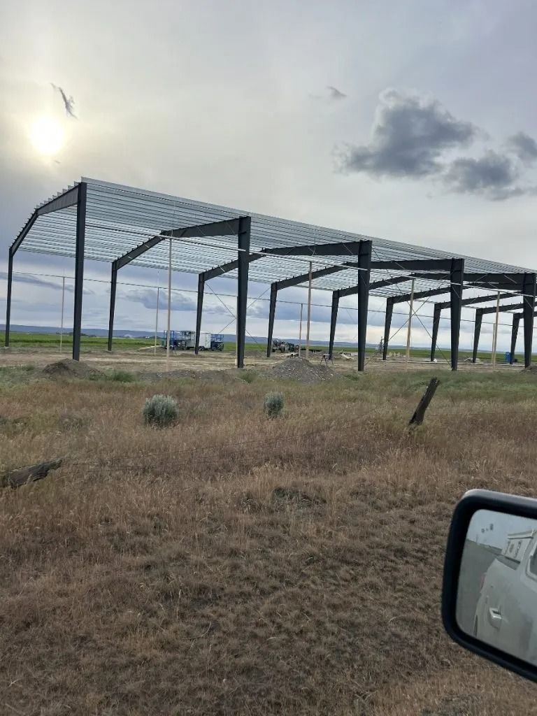 The steel frame of a large rectangular building stands in an open, grassy field under a cloudy sky.