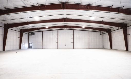 An interior view of a wide, empty industrial warehouse with steel frame beams and white walls.