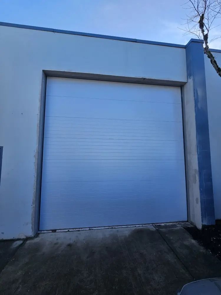 A closed white roll-up garage door centered on the exterior wall of a white building under a clear blue sky.