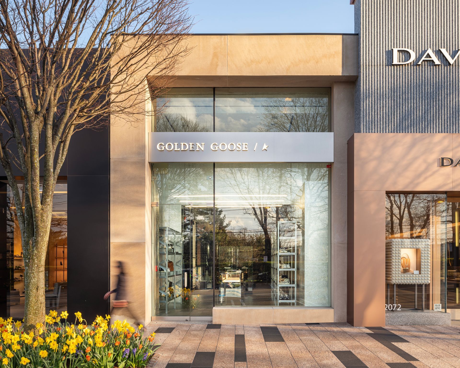 Golden Goose store facade with a glass front, surrounded by beige and brown buildings, and a person walking by.