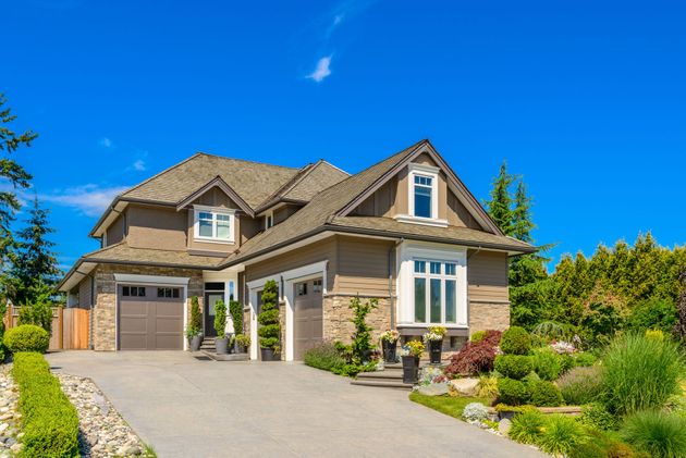 A two-story tan house with stone accents, a multi-gabled roof, a paved driveway, and lush landscaping under a blue sky.