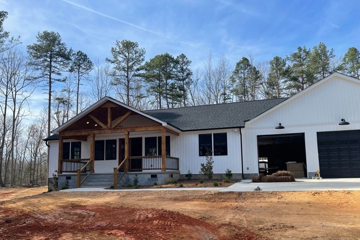A modern white farmhouse with a timber-framed front porch and attached garage, situated on a wooded lot with dirt ground.