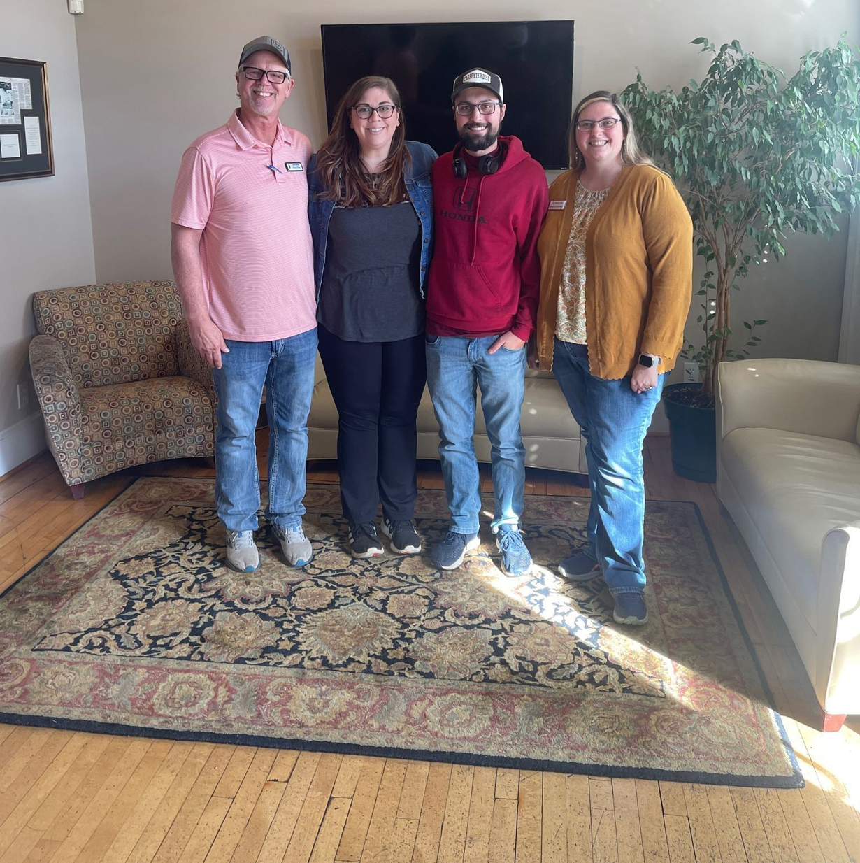 Four people stand smiling together in a room with a patterned rug, a television, and furniture.