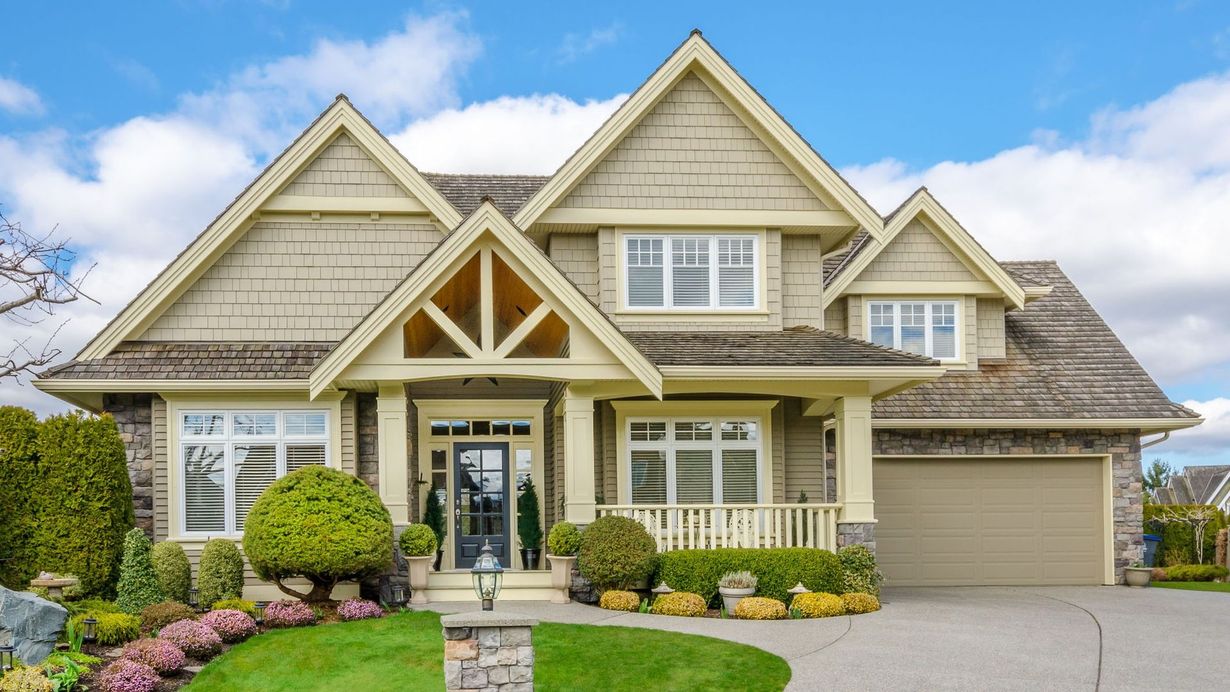 A two-story suburban home with tan shake siding, a stone facade, a covered porch, and an attached two-car garage.